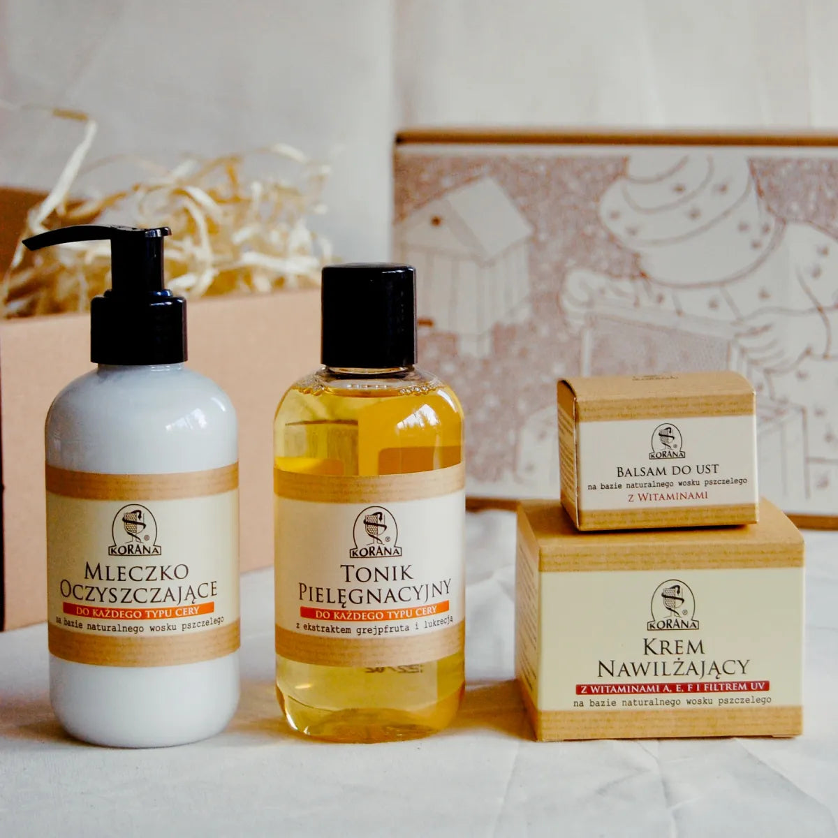 Mleczko oczyszczające na bazie wosku pszczelego, tonik pielęgnacyjny z ekstraktem grejpfruta i lukrecją, balsam do ust na bazie wosku pszczelego i krem nawilżający z witaminami i filtrem UV na bazie wosku pszczelego. Produkty stoją na tle ozdobnego opakowania na prezent